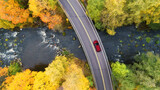 Fototapeta Na ścianę - Aerial view of road and bridge over river with red car in yellow and orange autumn forest in rural Finland.