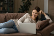 © fizkes - Interested millennial lady rest on couch with laptop read blog write comment to social network chat with mate online by wireless internet. Happy young female lying down on sofa working from home on pc