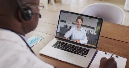 Poster - African american male doctor wearing phone headset taking notes while having a video call on laptop