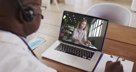 Wall Mural - African american male doctor wearing phone headset taking notes while having a video call on laptop