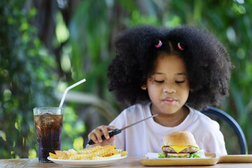 Sticker - childhood and eating concept - little african american curly hair girl holding knife and looking at burger and french fries on the table .enjoying unhealthy food.