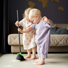 Cute Little Children Infant Boy And Toddler Girl In Linen Clothes Sweeping Floor In Room With Broom, Helping Mom With House Chore At Home. Baby Boy Brother And Older Sister Spending Time Together