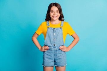 Canvas Print - Photo of shiny pretty small school girl wear denim jeans overall arms waist smiling isolated blue color background