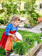 Wall Mural - Beautiful little toddler girl watering summer or spring flowers and herbs with kids bucket. Happy child helping in family garden, outdoors. Activity for children.