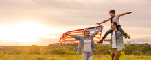 Young Parents With Their Daughter Holding American Flag In Countryside At Sunset. Independence Day Celebration