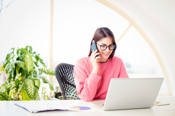 Young woman using mobile phone and laptop while sitting at desk and working from home