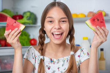 Wall Mural - Beautiful young teen girl holding big Watermelon while standing near open fridge in kitchen at home