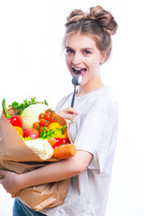 Smiling Caucasian Female Holding Eco Paper Bag Filled With Multiple Vegetables and Groceries Holding Spoon in Front of Mouth Against White Background.