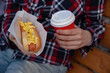 a hot dog and a red cup of coffee in the hands of an impersonal girl in a plaid shirt on a wooden bench
