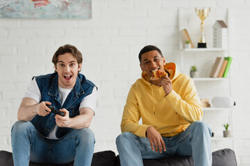 Wall Mural - KYIV, UKRAINE - MARCH 22, 2021: interracial friends playing video game and enjoying pizza on couch in modern living room