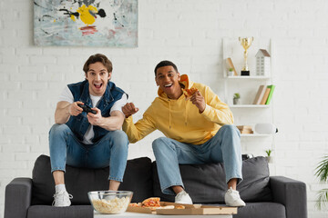 Wall Mural - KYIV, UKRAINE - MARCH 22, 2021: interracial friends playing video game with joystick and enjoying pizza and popcorn on couch in modern living room