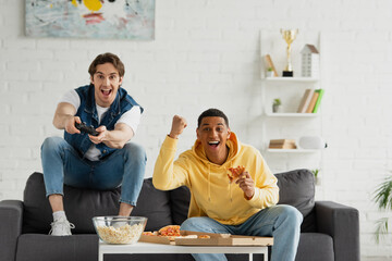 Wall Mural - KYIV, UKRAINE - MARCH 22, 2021: interracial friends playing video game with joystick and enjoying pizza on couch in modern living room