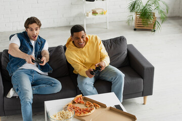 Wall Mural - KYIV, UKRAINE - MARCH 22, 2021: high angle view of interracial friends emotionally playing video game with joysticks and enjoying pizza on couch in modern living room