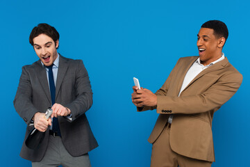 Wall Mural - smiling african american man filming with smartphone how his friend opening bottle of champagne isolated on blue