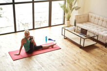 High Angle View Of Mature Woman Going In For Sports At Home, Sitting On Yoga Mat And Stretching Her Body In Front Of Laptop In Living Room