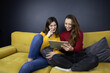 © Model Republic - two pretty long-haired brunette girls chatting and messaging sitting on sofa in stylish living room. Young ladies hold tablet listening music, and communicating on social networks. quarantine lockdown