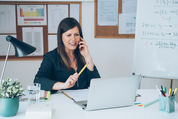Wall Mural - Female professor having break with online class and talking on mobile phone .
Cheerful teacher calling somebody on phone and preparing to working from home desk with E-learning students online.