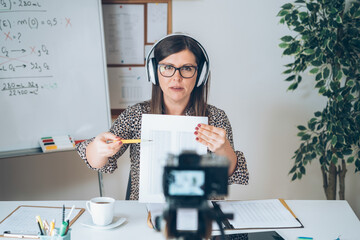 Wall Mural - Professor working at home, looking and talking direct to video camera and pointing to paper.
Teacher holding online class for e-learning students, recording math lesson using camera and headset.