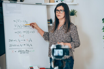 Wall Mural - Teacher recording video tutorial for e-learning class.
Female professor standing and explaining the assignment pointing on the whiteboard and using camera for live streaming online on the internet.
