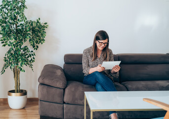 Wall Mural - Cheerful Business Woman Sitting on Sofa and using Tablet
Smiling businesswoman with wireless earphones looking in digital tablet, working at home concept.