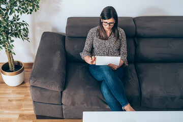 Wall Mural - Business Woman Sitting on Sofa and Using Tablet
High angle view of serious businesswoman with wireless earphones looking in digital tablet while sitting on couch in living room, working at home online