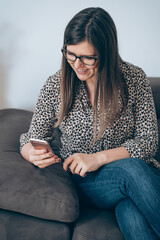Wall Mural - Cheerful Woman Sitting on Sofa and Using Mobile Phone at Home
Smiling businesswoman working at home, calling or typing message on smartphone while relaxing on couch in living room.