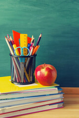 Back to school background. Pens, pencils, rulers, scissors in a glass, a red apple, a stack of notebooks and textbooks on the background of a green school board, closeup, copy space.