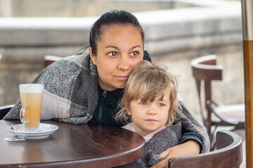 Street portrait of a mother and little daughter wrapped in blankets, sitting at a table in a summer cafe over a cup of coffee latte.