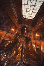 Gorgeous Brunette Girl In Haute Couture Dress Made Of Gold Feathers On The Background Of Luxurious Historical Palace Interiors.