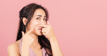 Portrait teenage Asian beautiful young woman having skin problems squeezing pimples on her face, studio shot on pink background, with copy space, Thai female acne, beauty care concept