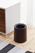 Subject shot of compact cylindric wastebasket with garbage bag in it. Brown trash can without lid is located on the wooden floor near drawer unit in light room.