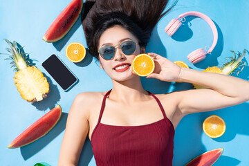 Beautiful asian woman in red bikini, sunglasses and lying on blue background with headphones, phone and fruits around. Tropical summer beach travel concept