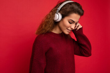 Closeup portrait of attractive positive young brunet curly woman wearing dark red sweater isolated over red background wall wearing white bluetooth headphones listening to music and having fun looking