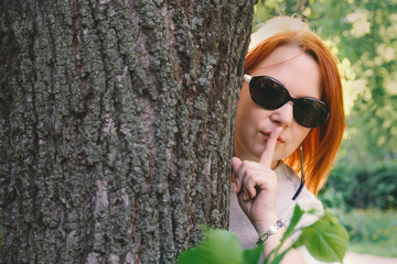 Young woman with red hair in sunglasses peeks out from behind a tree and shows gesture quietly