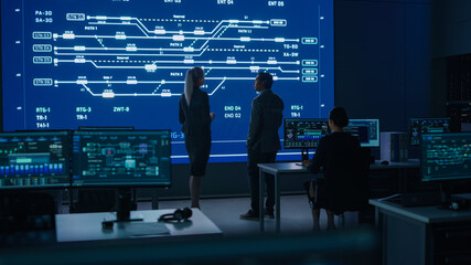 Project Manager and Computer Science Engineer Talking while Using Big Screen Display Showing Infrastructure Infographics and Data.Telecommunications Company System Control and Monitoring Room