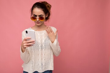 Photo of attractive dissatisfied young woman wearing casual white blouse and colorful sunglasses isolated over pink background wall holding and using phone looking at gadjet display