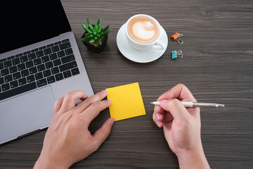 Wall Mural - Young man hand holding blank yellow sticky note on modern table desk, laptop, green plant, coffee cup, paper clip, text and massage with notepad