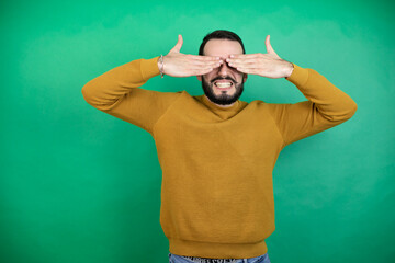 Wall Mural - Handsome man wearing casual clothes over isolated green background covering eyes with hands smiling cheerful and funny. Blind concept.