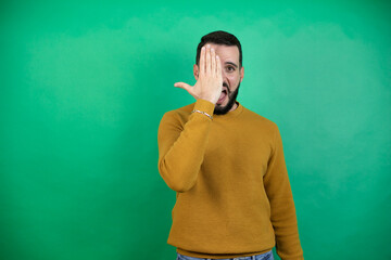 Wall Mural - Handsome man wearing casual clothes over isolated green background covering one eye with hand, confident smile on face and surprise emotion.