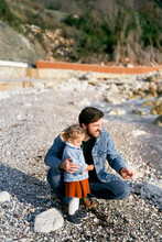Smiling Dad Squatted Next To The Little Girl, Handing Her Pebbles From A Rocky Beach By The Water. Close-up