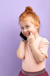 © alfa27 - Cute child holding cell phone, smiling, having nice talk with someone isolated on purple studio background. Redhead kid girl in casual t-shirt stand having conversation