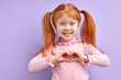 © alfa27 - portrait smiling happy little girl with red hair making heart sign with hands isolated on purple background. Positive human emotion expression feeling life perception body language