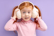 © alfa27 - Happy little girl with red hair stand wearing wireless headphones, enjoy listening to music, on purple studio background. child in pink shirt looking at camera with surprised facial expression