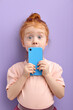 © alfa27 - surprised redhead child girl using smartphone, isolated on purple background in studio. checking news, surf the net, look at camera with big blue eyes, closing face with blue mobile phone