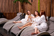 © alfa27 - three women in white bathrobes sitting on chaise-longue and drinking champagne at spa, celebrating, having conversation, clinking glasses