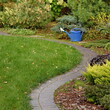 Backyard Garden Modern Design Landscaping. Decorative Garden Winding Pathway Walkway From Black Bricks. Back Yard Lawn And Natural Mulched Border Between Grass And Curved Brick Paving. Path To House.