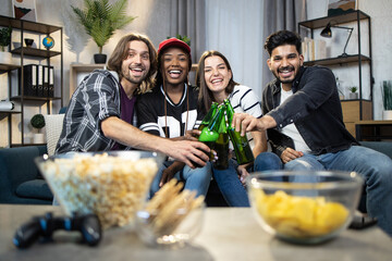 Wall Mural - Group of happy multicultural friends sitting together on couch drinking beer and eating snacks. Cheerful young people enjoying carefree weekends at home.