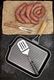 Fototapeta Desenie - Raw Stuffed Sausages, Empty Grill Pan and Grill Tools On Rustic Black Table Background, Top View. Raw Sausages In Natural Casing. Sausages For Grilling or Frying On Paper Overhead View.