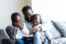 Father Reads A Book To His Two Daughters On Sofa Whilst They Laugh