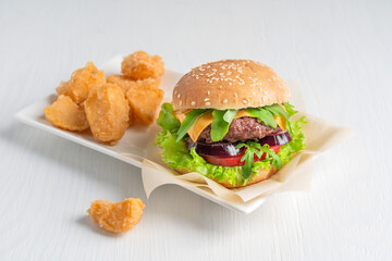 Wall Mural - Organic juicy burger made of grilled beef meat, cheese, lettuce, onion and tomatoes inside sliced bun with sesame seeds served with fried potato wedges on rectangular plate on white wooden table